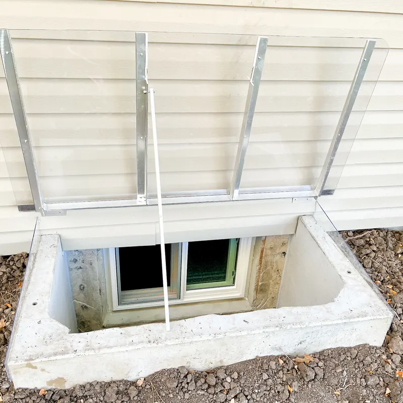 Window well cover propped open with support rod, showing clear polycarbonate and aluminum frame